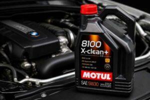 Kanister oleju Motul 8100 X-clean+ 5W-30 ustawiony na tle silnika samochodowego